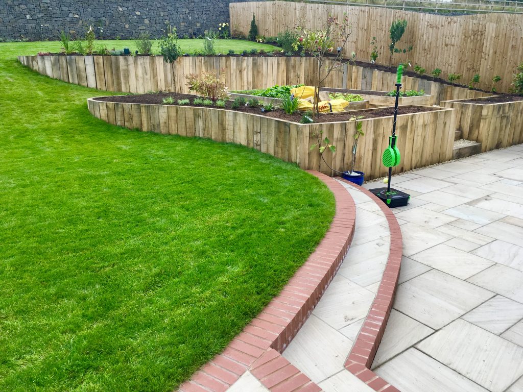 Edging and detailing in landscape gardens depicted by this image of a patio meeting a raised lawn with a long, sweeping curve divided with a brick edging detail to the steps rising to it.